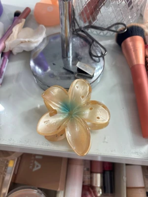 Blommig hårklämma i pärlemor - Säljer en hårklämma formad som en blomma i skimrande pärlemor med en svag blå ton i mitten. Klämman är gjord i plast och har en mjuk, glansig finish. Perfekt för att piffa upp frisyren med en söt detalj.
