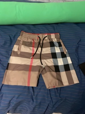 Rutig shorts från Burberry - Säljer ett par klassiska rutig shorts med beige, svart, vit och röd färg. Shortsen har dragsko i midjan och sidofickor. Perfekta för sommaren. OBS! STORLEK XS MEN KAN PASSA S. 
