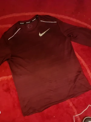 Vinröd långärmad Nike Dri-Fit tröja - Snygg vinröd långärmad träningströja från Nike med Dri-Fit material som andas. Tröjan har vita detaljer på axlarna och en silverfärgad Nike-logga på bröstet. Perfekt för löpning eller annan träning.