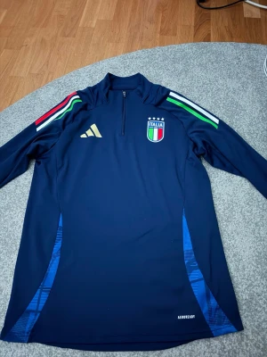 Italien långärmad träningsdress Adidas - Mörkblå långärmad träningströja från Adidas med Italiens landslagsmärke på bröstet. Tröjan har halv dragkedja, guldigt Adidas-logga, samt ränder i grönt, vitt och rött på axlarna. Tillverkad i funktionsmaterial Aeroready. Det är en komplett dress identiska mjukis byxor i samma material tillkommer dressen är i helt nytt skick och bara använt vid max 5 tillfällen där jag har tränat. OBS överdelen är i storlek L och byxorna är i storlek M. (Pris är såklart inte hugget i sten)
