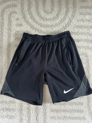 Svarta Nike Shorts - Säljer ett par svarta träningsshorts från Nike med elastisk midja och dragkedjefickor på sidorna. Shortsen har gråa detaljer vid benen och den klassiska Nike-loggan i vitt på vänster ben. Perfekta för sport eller gym!
