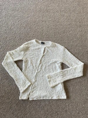 Vit crinklad topp med knappar - Säljer en tunn, vit topp med crinklad struktur och rund halsringning. Koftan har långa ärmar och stängs med små knappar framtill. 
