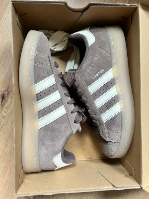 Adidas Campus bruna sneakers - Säljer ett par Adidas Campus sneakers i brun mocka med klassiska vita ränder på sidorna och vit detalj vid hälen. Skorna har en låg profil och gummisula som ger en clean och retro vibe. Perfekta för dig som gillar stilrena och tidlösa sneakers. Använda endast en gång, säljer för att de är lite för små. Nypris är 1400kr. Storleken är 39 1/3. Pris går att diskuteras. Är även öppen för att byta skor om man har nästan lika fast i 40 eller 41.