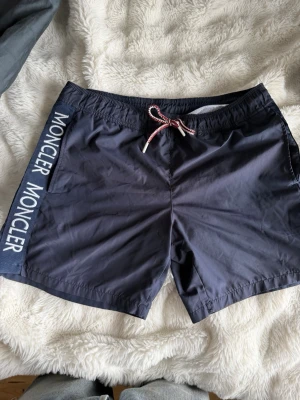 Mörkblå badbyxor från Moncler - Snygga mörkblå badbyxor från Moncler med vit logotyp längs ena sidan och rödvit dragsko i midjan. Klassisk passform med elastisk midja och bakficka med Moncler-märke. Perfekta för stranden eller poolen.