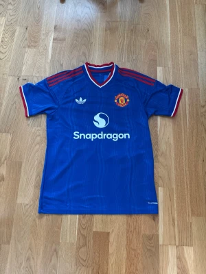 Blå Manchester United matchtröja - Säljer en blå Manchester United matchtröja med röd och vit krage och röda detaljer på axlarna. Tröjan har klubbmärke, Adidas-logga och Snapdragon-tryck på framsidan samt Mainoo och nummer 37 på ryggen. Perfekt för fotbollsfans!