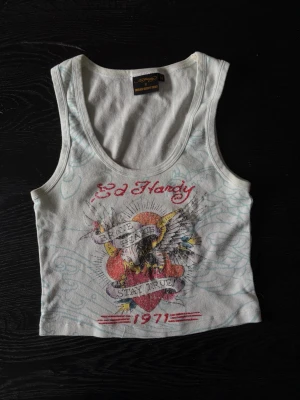 Vit Ed Hardy linnetopp - Säljer en vit linnetopp från Ed Hardy med färgglatt tryck framtill som visar en örn, hjärta och texten 'Brave Heart Stay True 1971'. 