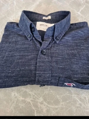 Mörkblå skjorta från Hollister storlek S/M - Snygg mörkblå skjorta från Hollister med diskret melerad look och klassisk krage. Skjortan har knappstängning framtill och en bröstficka med Hollisters logga broderad i vitt och rött. Perfekt till jeans eller chinos. Den passar både S & M.