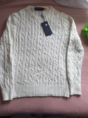 polo ralph lauren tröja vit - Kabelstickad vit tröja från Polo Ralph Lauren med rund halsringning och ribbade muddar. Klassisk och stilren design som passar till det mesta. Tillverkad i mjukt material för en skön känsla.