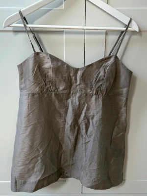 Beige linnetopp med smala axelband - Snygg beige linnetopp med smala axelband och lätt figurnära passform. Toppen har en enkel och stilren design med söm under bysten för en feminin siluett. Perfekt att matcha med jeans eller kjol för en avslappnad look. HELT NY