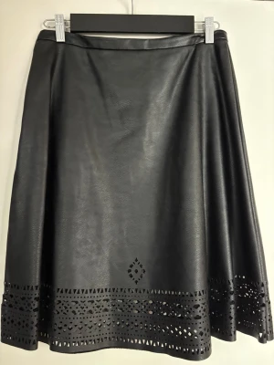 Svart skinnkjol från BOSS - Snygg svart kjol i eco-läder från BOSS. Kjolen har A-linjeformad passform och dekorativa laserskurna detaljer längs nederkanten. Perfekt för dig som vill ha en edgy och trendig look. Materialet känns mjukt och följsamt.