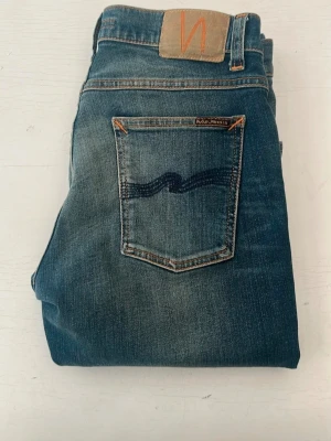 Blå jeans från Nudie Jeans - Snygga blå jeans från Nudie Jeans med klassisk femficksmodell och orangea sömmar. Jeansen har en diskret tvättad look och vågig brodyr på bakfickorna. Tillverkade i denim med en bekväm passform och normal midja. Modellen är Tight Terry, kom gärna med bud!