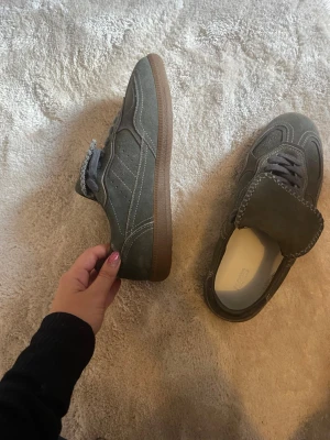 Grå sneakers i mocka med gummisula - Säljer ett par grå/gröna sneakers i mocka med vita sömmar och klassisk gummisula. Skorna har lågt skaft och snörning framtill. Perfekta för dig som gillar stilrena och tidlösa sneakers med en touch av retro. Skorna är från Aloha och ny pris ligger på drygt 2500kr 