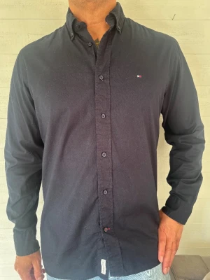 Tommy Hilfiger skjorta - Klassisk svart skjorta från Tommy Hilfiger med knappslå och liten logga på bröstet. Skjortan har lång ärm, button down-krage och är tillverkad i ett skönt material. Perfekt för en clean och stilren look.