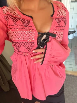 Rosa odd Molly blus med broderi och rosett - Supersöt rosa blus i boho-stil med broderade detaljer i svart och vitt över bröstet. Blusen har en djup urringning med svart rosettdetalj och vågiga kanter. Långärmad modell i lätt och luftig bomull, perfekt för en chill och trendig look.