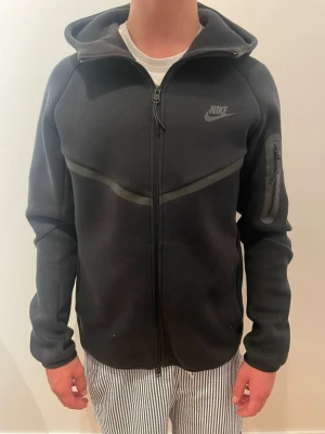 Svart Nike tech hoodie  - Säljer en svart hoodie från Nike med hel dragkedja framtill och huva. Tröjan har en diskret Nike-logga på bröstet och en praktisk ficka med dragkedja på ena ärmen. Tillverkad i mjukt material och har en sportig look med svarta detaljer.