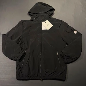 Helt ny Moncler tunnare jacka med luva - Strl L - Säljer en superfint tunn jacka från Moncler med snygg luva. Jackan är i klassiskt svart utförande och är helt ny och oanvänd med alla lappar (tags) kvar. Den perfekta vår- eller höstjackan som passar till allt! Storlek: L (PASSAR PERFEKT FÖR M) Skick: 10/10 (oanvänd, se bilder för     detaljer)