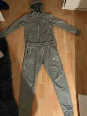 Grått Nike loungewear set - Mysigt grått loungewear set från Nike med dragkedja på hoodien och justerbar midja på byxorna. Setet har huva, fickor på både överdel och byxor samt Nike-logga på bröstet och benet. Perfekt för chill dagar hemma.