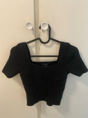 Svart croppad topp med fyrkantig ringning - Trendig svart croppad topp med fyrkantig ringning och korta ärmar. Toppen är ribbad och har en tight passform som framhäver figuren. Perfekt att matcha med höga jeans eller kjol för en snygg look.