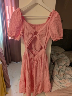 Rosa kortklänning med cut-out och puffärm - Söt rosa kortklänning med puffärmar. Klänningen har en ringformad detalj vid bysten, smockad rygg och volang i kjolen. Perfekt för sommarens alla tillfällen.och ger en romantisk vibe. Storlek 158 men jag skulle säga att den är i storlek S men passar många eftersom att den är så stretching.
