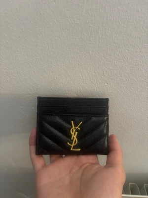 YSL plånbok/korthållare - Snyggt svart kortfodral i läder från Yves Saint Laurent med guldfärgad YSL-logga framtill. Fodralet har quiltad design och flera fack för kort. Perfekt accessoar för dig som vill ha något stilrent och lyxigt i fickan.