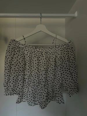 Vit prickig off shoulder-blus - Söt vit blus med svarta prickar och off shoulder-modell. Blusen har trekvartsärmar med resår vid ärmslut och midja, samt tunna axelband. 