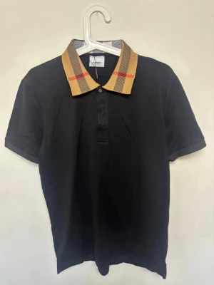 Svart pikétröja från Burberry - Snygg svart pikétröja från Burberry med klassisk beige och röd rutig krage. Tröjan har korta ärmar och knappar framtill. Perfekt för dig som vill ha en stilren men ändå unik topp med ikonisk Burberry-detalj.