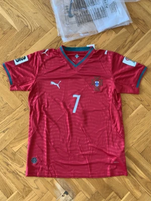 Portugal matchtröja Ronaldo #7 - Snygg röd Portugal matchtröja från Puma med Ronaldo och nummer 7 på ryggen. Tröjan har vita detaljer, vågmönster och gröna inslag vid krage och ärmslut. Perfekt för dig som älskar fotboll och vill stötta Portugal på match eller träning.
