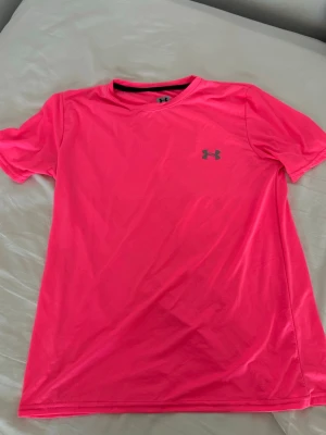 Neonrosa tränings-tshirt Under Armour - Snygg neonrosa tränings-tshirt från Under Armour med rund hals och korta ärmar. Tillverkad i ett lätt och ventilerande funktionsmaterial som passar perfekt för träning och sport. Diskret logga på bröstet.