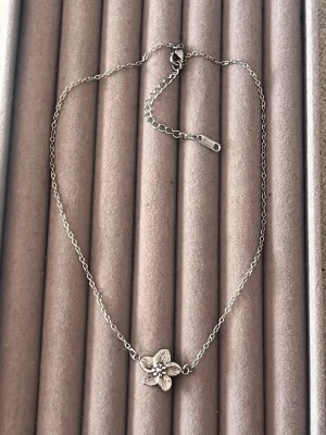 Silverfärgat halsband med blomma - Snyggt halsband i silverfärg med en detaljrik blomma som hänge. Kedjan är tunn och har justerbar längd med karbinlås. Perfekt för dig som gillar stilrena och feminina accessoarer. Materialet ser ut att vara metall.