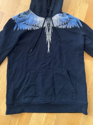 Svart Marcelo burlon hoodie  - Säljer en svart hoodie med stor huva och känguruficka. Runt axlarna och nacken finns ett detaljerat tryck av blå och vita vingar som fortsätter över ryggen. Hoodien har dragsko i huvan och är gjord i mjukt bomullstyg. Perfekt för dig som vill sticka ut med en unik design.