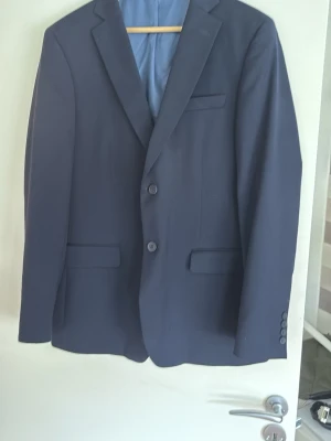 Mörkblå klassisk kavaj herr - Snygg mörkblå kavaj med två knappar framtill, klassisk passform och slät finish. Kavajen har två fickor med lock, bröstficka och knappar vid ärmslut. Perfekt till skjorta och jeans för en stilren look. Använts 1 gång, säljer pågrund av att jag fått en ny. Kavajstorlek: 52 regular slimfit. Byxstorlek: 34/32 slimfit.