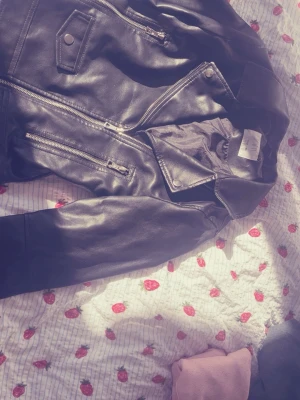 Svart klassisk skinnjacka - Snygg svart skinnjacka med flera dragkedjor och coola detaljer. Jackan har bred krage, fickor med dragkedja och tryckknappar. Perfekt för dig som vill ha en tidlös bikerstil och matcha med jeans eller kjol.