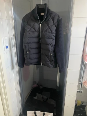 Svart cardiganjacka från Moncler - Snygg svart cardiganjacka från Moncler med quiltad front och ribbstickade ärmar. Jackan har hög krage, två fickor med tryckknappar och dragkedja framtill. Moncler-logga på ärmen ger en exklusiv touch. Perfekt för dig som vill ha både stil och komfort. Sällsynt med black label. Size 1 motsvarar S