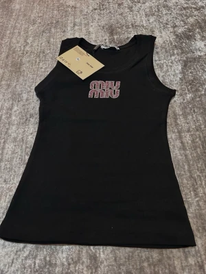 Svart ribbad linnetopp Miu Miu - Snygg svart ribbad linnetopp från Miu Miu med glittrande logga framtill. Toppen är ärmlös och har en tight passform som framhäver figuren. Perfekt att styla med jeans eller kjol för en trendig look.