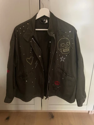 Grön jacka  - Cool olivgrön vindjacka med dragkedja och krage. Jackan har broderade detaljer som döskalle, hjärta, stjärnor och peace-märke samt små nitar. Två stora fickor framtill och patch på ryggen. Perfekt för dig som gillar unik streetstyle.