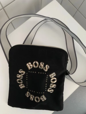 Svart axelväska från Hugo Boss - Svart axelväska i tyg från Hugo Boss med rundad fyrkantig form. Väskan har ett tryck med texten 'BOSS' i silver och 'HUGO BOSS' i mindre bokstäver. Justerbar axelrem i grått och vitt. 