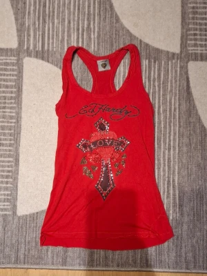 Röd Ed Hardy linnetopp med tryck - Snygg röd linnetopp från Ed Hardy med stort glittrigt tryck av ett kors och texten 'LOVE' framtill. Toppen har brottarrygg och ärmlös design, perfekt för dig som vill sticka ut med en edgy look.