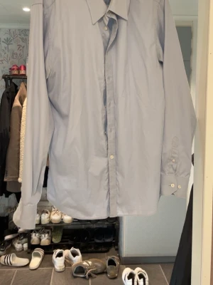 Ljusblå klassisk skjorta - Säljer en ljusblå skjorta med lång ärm och klassisk krage. Skjortan har knappar framtill och vid ärmsluten. Passar perfekt till både jeans och chinos för en clean och stilren look. Materialet ser ut att vara bomull eller en bomullsblandning.