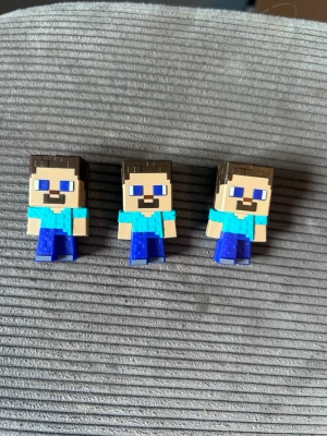 Minecraft Steve minifigurer 3-pack - Tre små Minecraft Steve-figurer i plast med fyrkantig form, blå byxor och ljusblå tröja. Perfekta som samlarprylar eller dekoration till skrivbordet. Figurerna har brunt hår och skägg samt tydliga pixlade detaljer.