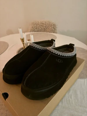 Svarta UGG tofflor med mönstrad kant - Säljer ett par svarta UGG tofflor i mocka med fluffigt foder och en snygg vitmönstrad kant runt öppningen. Tofflorna har en chunky sula och klassisk slip-on design som är både bekväm och trendig. Dom är lite nedtryckta i fluffet på ena skon men inget som känns när man har foten i 