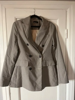 Grå dubbelknäppt kavaj Gina Tricot - Stilren grå kavaj från Gina Tricot med dubbelknäppning och svarta knappar. Kavajen har klassisk krage, två fickor med lock och en bröstficka. Materialet är strukturerat och fodret är ljust. Perfekt för en trendig och snygg look.