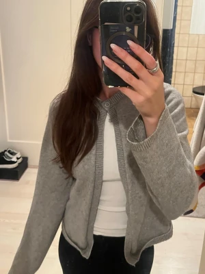 Ljusgrå kofta med knapp vid hals - En ljusgrå kofta från gina med rund halsringning och ribbade muddar. Koftan har en knapp uppe vid halsen som man kan knäppa om man vill, perfekt att slänga över en t-shirt. Koftan är i ttorlek S men skulle säga den passa XS också. I bra skick:)