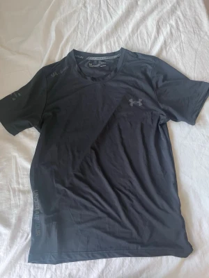 Svart tränings-tshirt Under Armour - Snygg svart tränings-tshirt från Under Armour med diskret logga på bröstet och tryck på sidan. Tillverkad i ett lätt och snabbtorkande funktionsmaterial som passar perfekt till gymmet eller löprundan. Klassisk rund hals och korta ärmar.