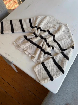 Vit stickad tröja med svarta ränder - Säljer en vit stickad tröja med breda svarta horisontella ränder. Tröjan har lång ärm, rund halsringning och en snygg rynkning i midjan som ger en cool siluett. Materialet är ribbstickat och känns mjukt och bekvämt. Den har några lite synliga fläckar på backsidan och en blå/grön fläck vid slutet av ärmen. 