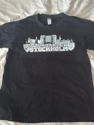 Svart Djurgården Stockholm t-shirt - Säljer en svart t-shirt med tryck av Djurgården Stockholm och stadssilhuett på bröstet. T-shirten har klassisk passform och korta ärmar. Perfekt för dig som vill visa ditt stöd för Djurgården på ett stilrent sätt. Passar om man är 170
