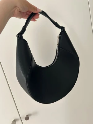Svart halvmåneformad handväska Mango - Stilren svart handväska i halvmåneform från Mango med kort axelrem och silverfärgad dragkedja. Väskan är gjord i slätt konstläder och har en modern, minimalistisk look som passar till många outfits.