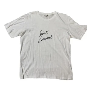 Saint Laurent t-shirt - Säljer en klassisk vit t-shirt från Saint Laurent med svart handskriven logga på bröstet. T-shirten har rund halsringning och korta ärmar. Perfekt basplagg för en clean och stilren look.
