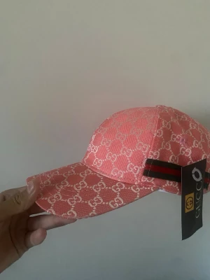 Rosa keps  med GG-mönster - Snygg rosa keps med klassiskt GG-mönster över hela kepsen. Kepsen har böjd skärm och justerbart band bak med röd-gröna ränder. Materialet är textil och kepsen har en modern streetstyle-vibe.