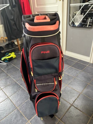 Svart golfbag från PING med röda detaljer - Snygg svart golfbag från PING med röda och gula detaljer. Flera rymliga fack med dragkedjor och praktiska fickor för tillbehör. Robust handtag upptill och vadderade axelremmar för enkel transport. Perfekt för dig som vill ha ordning på golfprylarna.