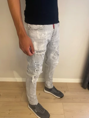 Ljusa slitna jeans med hål - Säljer ett par ljusgrå jeans från Dsquared2 med slitningar, hål och färgstänk för en riktigt trendig och avslappnad look. Jeansen har rak passform, klassiska fem fickor och patch med logga bak i midjan. Perfekta för dig som gillar streetstyle.